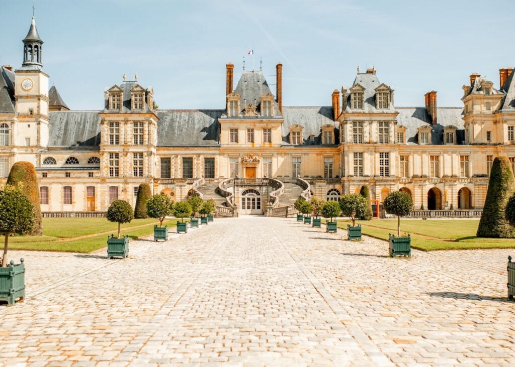 The courtyard of Château de Fontainebleau on a sunny day with the famous horseshoe staircase leading to the main entrance and potted topiaries lining the path