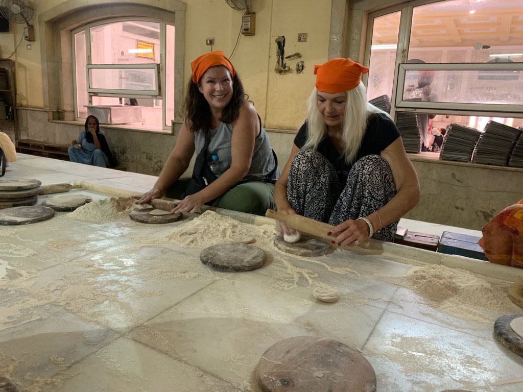 Two women in orange head coverings rolling bread dough on a flour covered table in a Sikh temple community kitchen in Delhi.