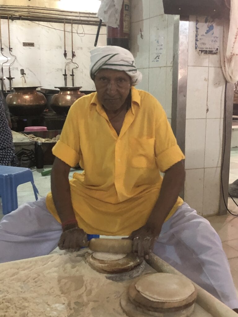 A man in a yellow shirt rolling dough in the community kitchen of a Sikh temple in Delhi, with large copper pots in the background.