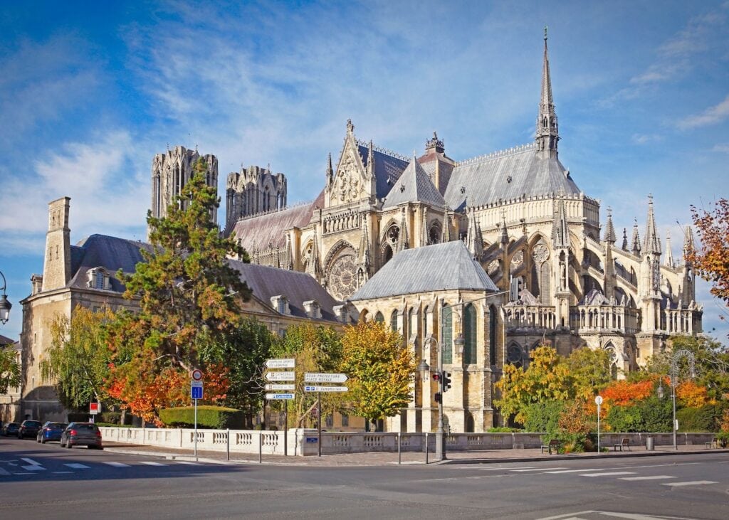 The Gothic exterior of the Cathédrale Notre-Dame de Reims in autumn with its flying buttresses and spires rising above colorful trees