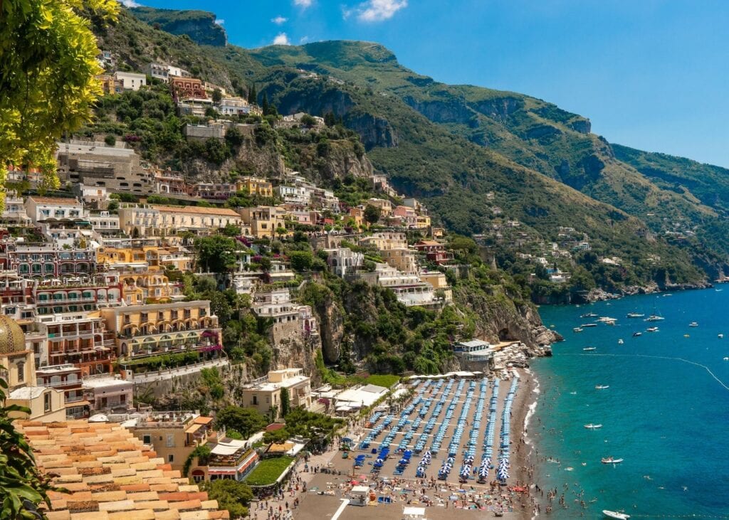 The pastel cliffside houses of Positano rising above a beach lined with blue umbrellas on Italy's Amalfi Coast.