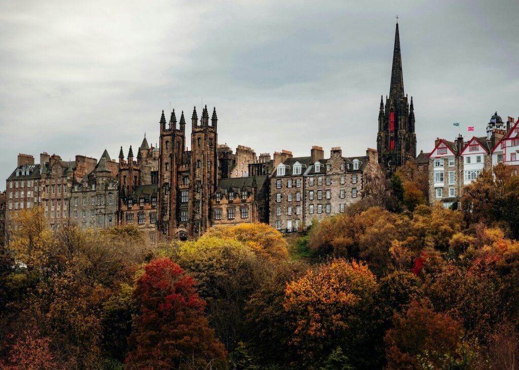 The spires and stone buildings of Edinburgh's Old Town rising above trees turning red and gold in autumn.