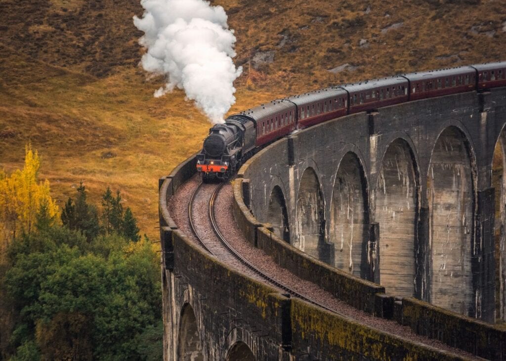 A steam train crossing the curved Glenfinnan Viaduct through the Scottish Highlands, with autumn hillsides in the background.