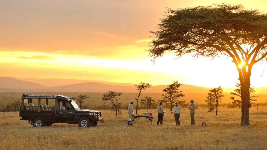 Safari vehicle parked near an acacia tree at sunset with guests enjoying sundowners on the savanna