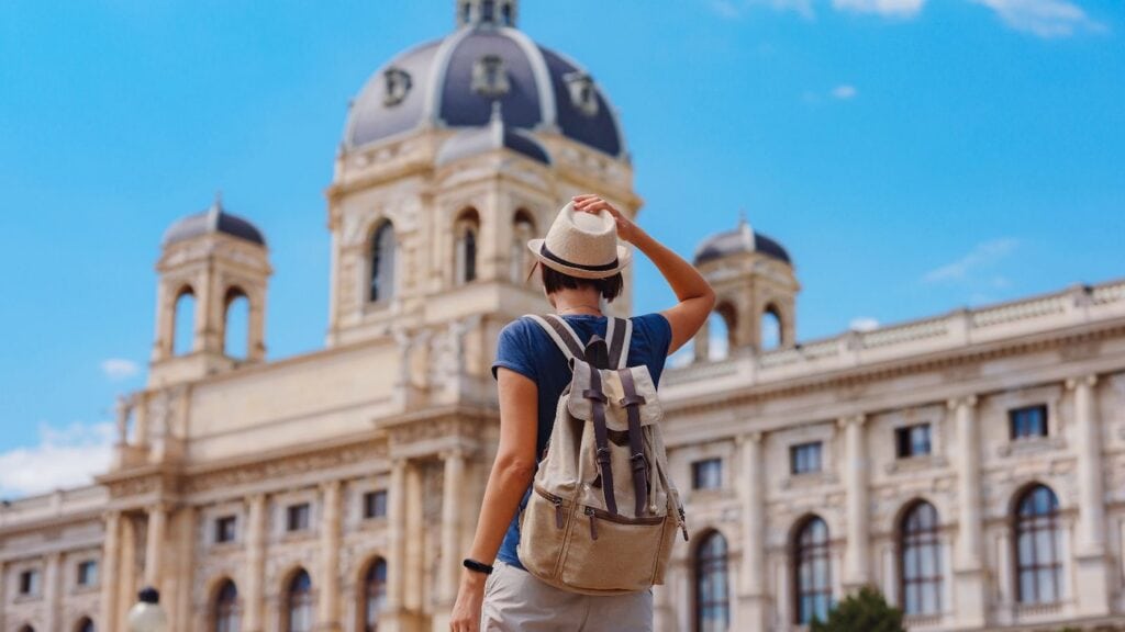 Solo female traveler with a backpack and hat standing in front of a grand European domed building on a sunny day.