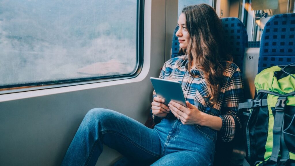 Solo female traveler sitting on a train with a backpack beside her, holding a tablet and looking out the window at the passing landscape.