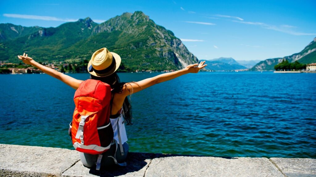 Woman with a red backpack and hat sitting at a lakeside with arms outstretched, surrounded by mountains and blue water on a solo trip.