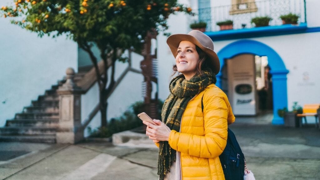 Woman in a yellow jacket and hat exploring a colorful European street solo, holding a phone and looking up with a smile.