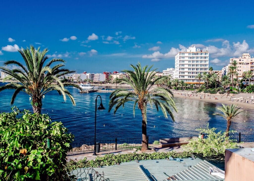 Palm tree lined waterfront in Ibiza Town Spain with a sandy beach, calm blue water, and hotel buildings along the shore