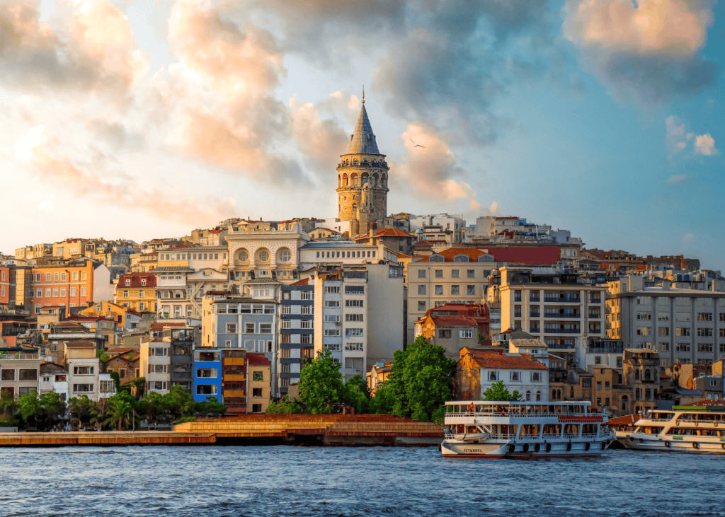 Galata Tower rising above the colorful hillside neighborhood of Karakoy in Istanbul Turkey with ferries on the Bosphorus in the foreground at golden hour