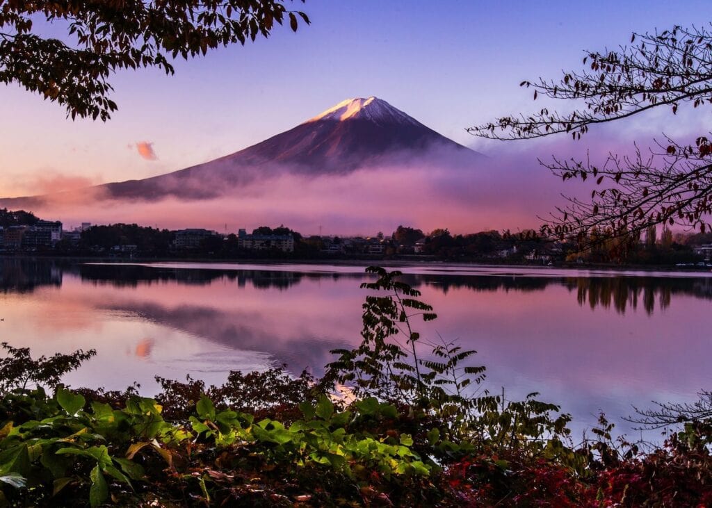 Mount Fuji rising above morning mist at sunrise with a glowing pink and purple sky reflected in a still lake surrounded by autumn foliage in Japan