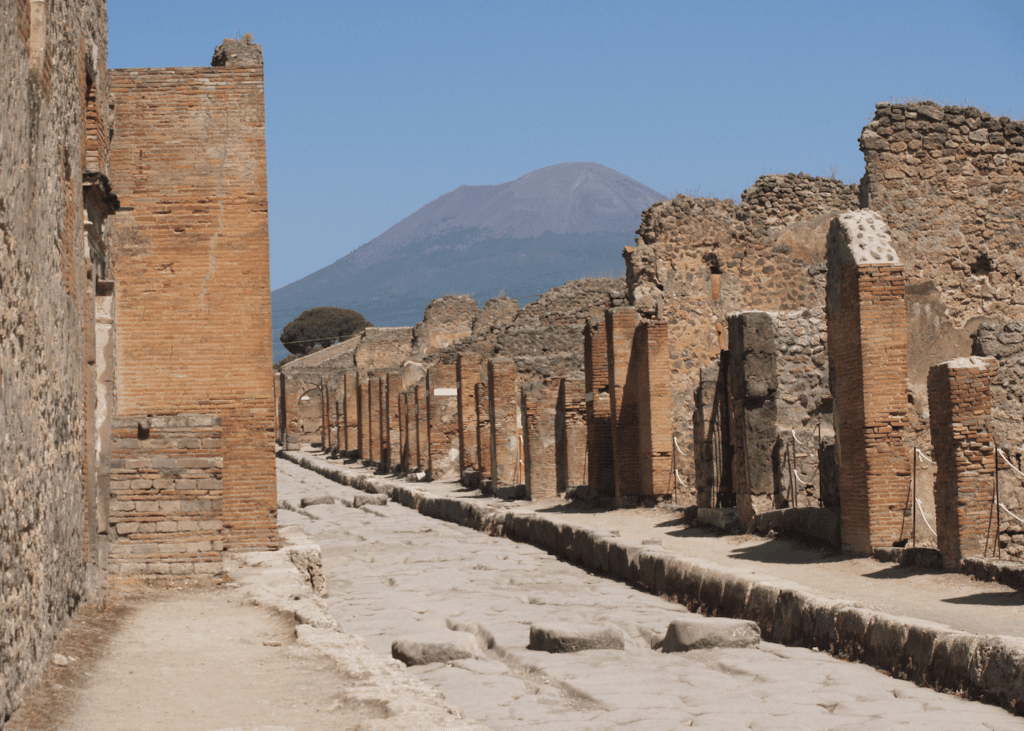 Ancient cobblestone street lined with brick ruins at Pompeii Italy with Mount Vesuvius visible in the distance under a clear blue sky.