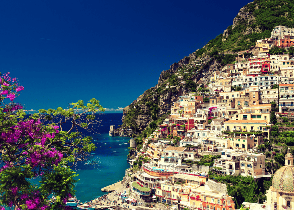 Colorful hillside buildings of Positano cascading down the cliffs above the turquoise water of the Amalfi Coast Italy with pink flowering trees in the foreground.