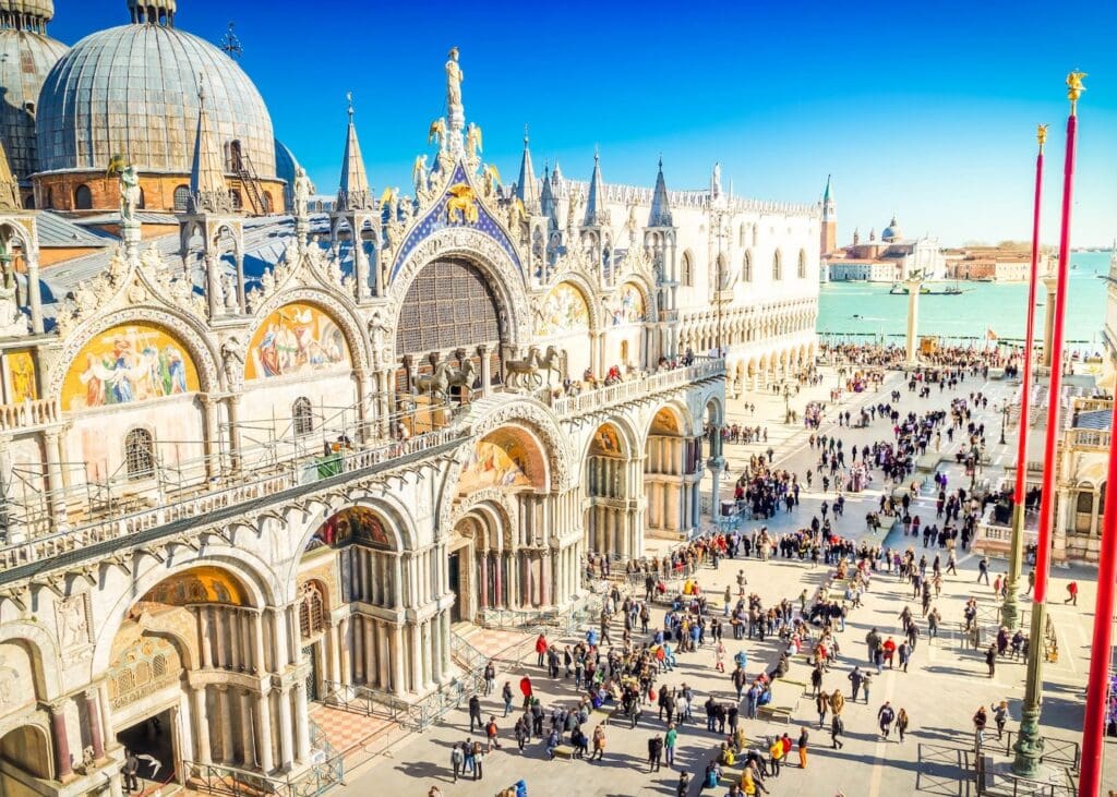 Aerial view of crowds of tourists filling St. Mark's Square in Venice Italy in front of the ornate gold mosaic facade of St. Mark's Basilica on a sunny day.