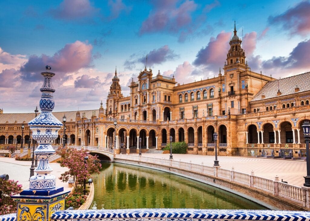 The ornate tiled facade and arched colonnade of Plaza de España in Seville Spain reflected in the surrounding canal under a dramatic cloudy sky.